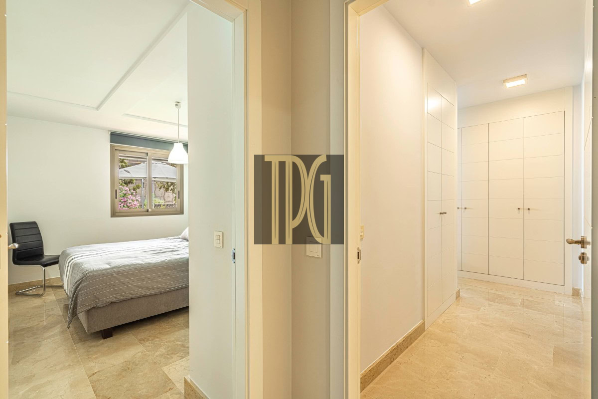 Bright hallway of a modern apartment leading to a bedroom on the left, which contains a bed, a black chair, and a window with greenery outside; white built-in closets line the right wall.