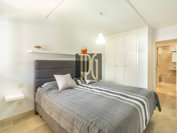 Modern bedroom with a gray upholstered bed, striped bedding, white walls, and a large built-in wardrobe; an open door reveals a beige-tiled bathroom.