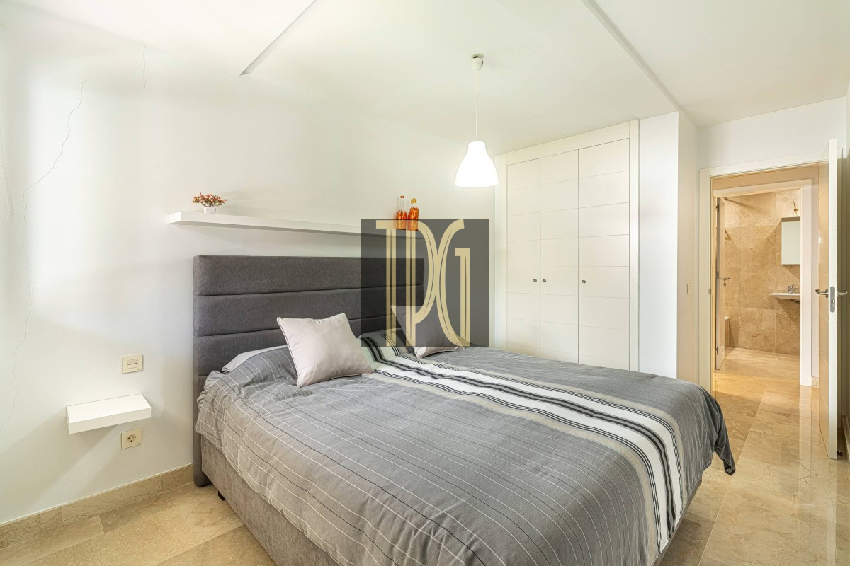 Modern bedroom with a gray upholstered bed, striped bedding, white walls, and a large built-in wardrobe; an open door reveals a beige-tiled bathroom.