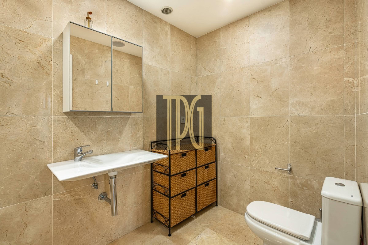 Beige tiled bathroom with a wall-mounted mirror cabinet, white sink, wicker storage drawers, and a toilet beside it.