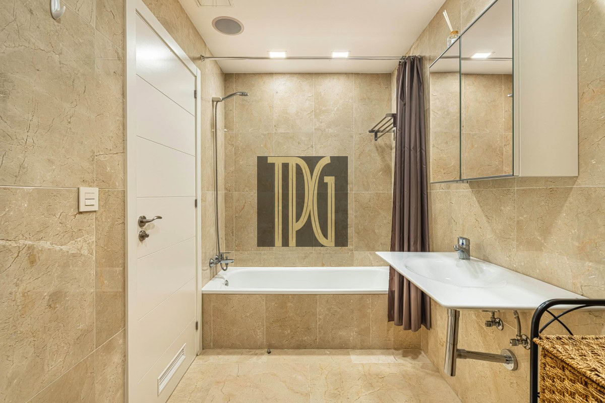 Modern beige-tiled bathroom with a white bathtub, shower head, gray shower curtain, and a white sink with a mirrored cabinet on the right.