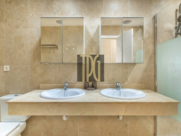 Beige marble bathroom with a double sink vanity, two oval sinks, and a glass-walled shower on the right.