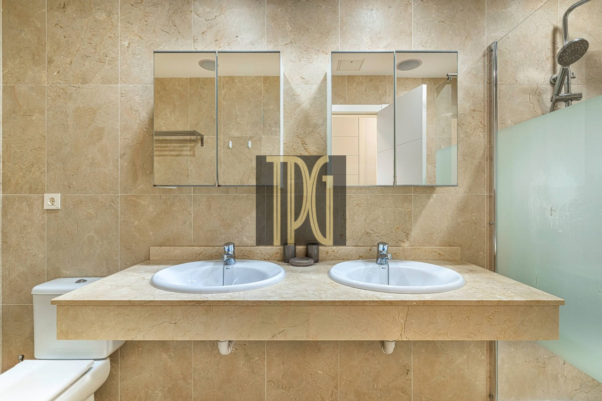 Beige marble bathroom with a double sink vanity, two oval sinks, and a glass-walled shower on the right.