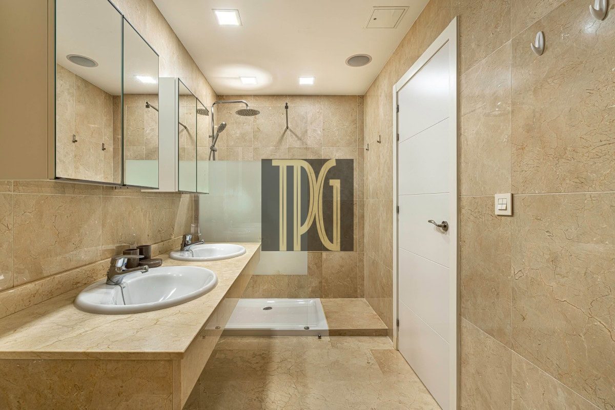 Bathroom with two oval sinks, beige marble countertop, mirrored cabinets, glass shower partition, and a white door in the background.