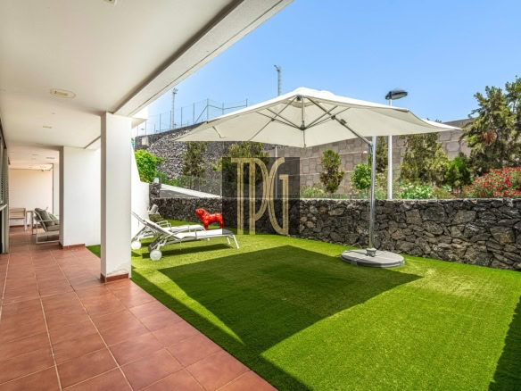 Patio with terracotta tiles along a white building, adjoining a green lawn and white lounge chairs under a large umbrella by a stone wall.