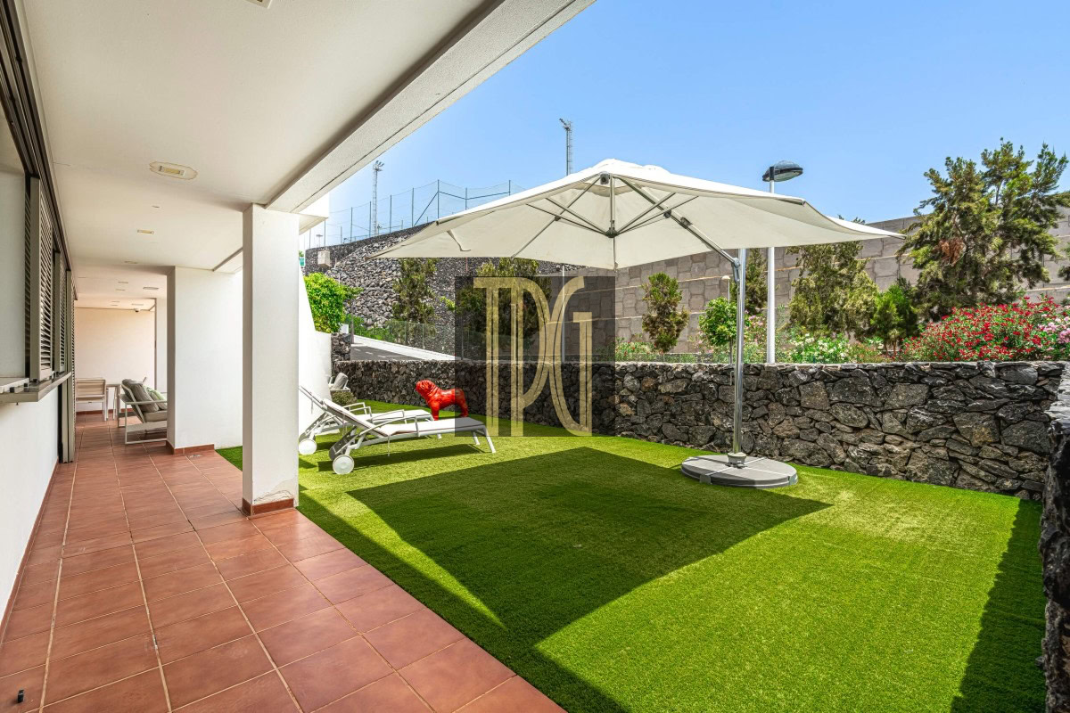 Patio with terracotta tiles along a white building, adjoining a green lawn and white lounge chairs under a large umbrella by a stone wall.