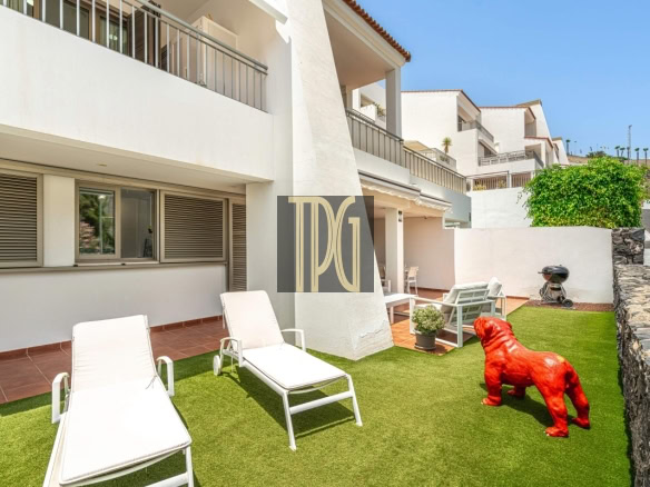 Patio of a white multi-story building with sun loungers on artificial grass and a red bulldog statue near a stone wall.