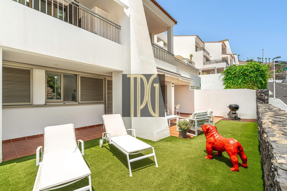 Patio of a white multi-story building with sun loungers on artificial grass and a red bulldog statue near a stone wall.