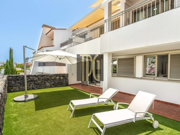 Outdoor patio of a modern apartment building with two white sun loungers on artificial green grass, a large umbrella, and a stone privacy wall nearby.