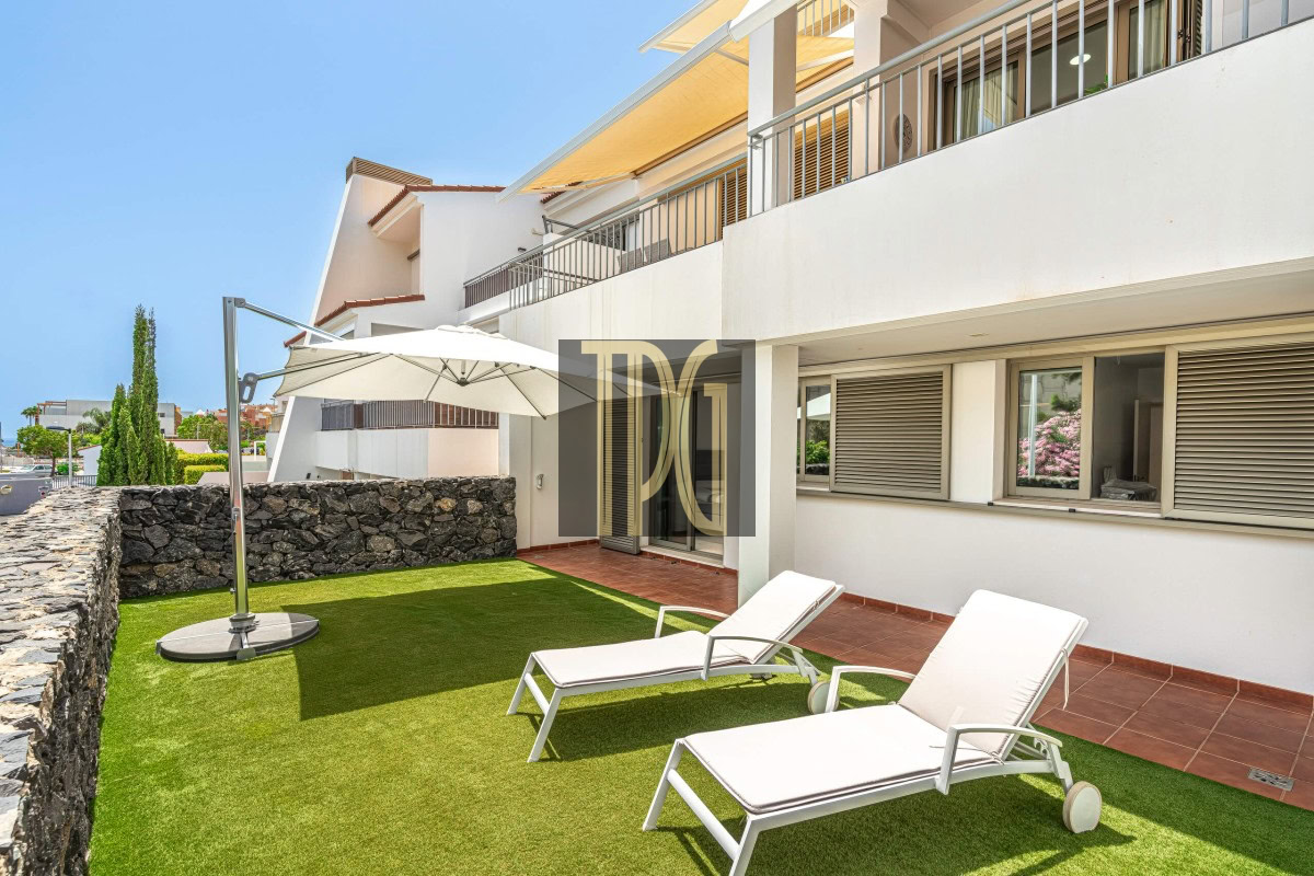 Outdoor patio of a modern apartment building with two white sun loungers on artificial green grass, a large umbrella, and a stone privacy wall nearby.