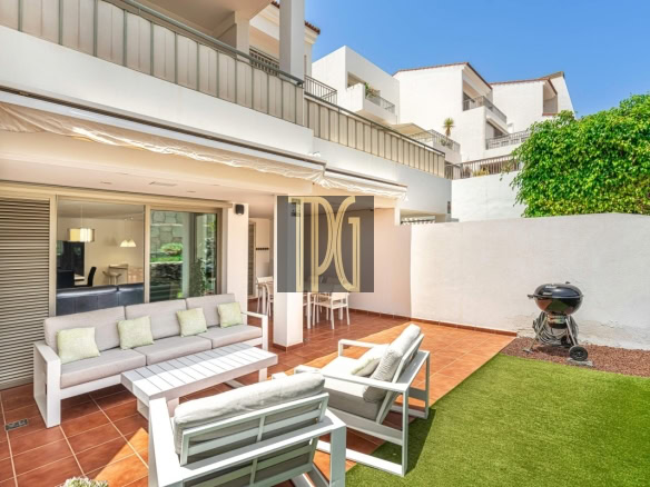 Sunny outdoor terrace of a modern white apartment building with a beige sofa set, white chairs, and a coffee table on terracotta tiles; a grill and green lawn are to the side.