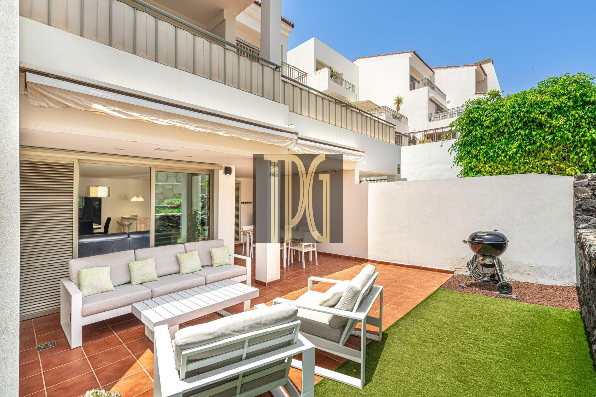 Sunny outdoor terrace of a modern white apartment building with a beige sofa set, white chairs, and a coffee table on terracotta tiles; a grill and green lawn are to the side.