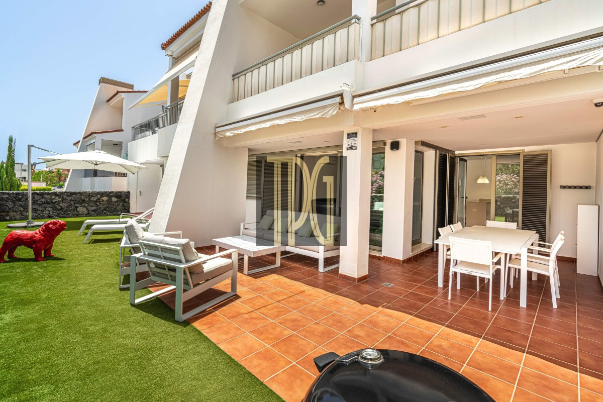 Outdoor patio of a white villa with terracotta tiles, white seating, and a dining table under a covered area; a small red statue dog stands on the lawn to the left.
