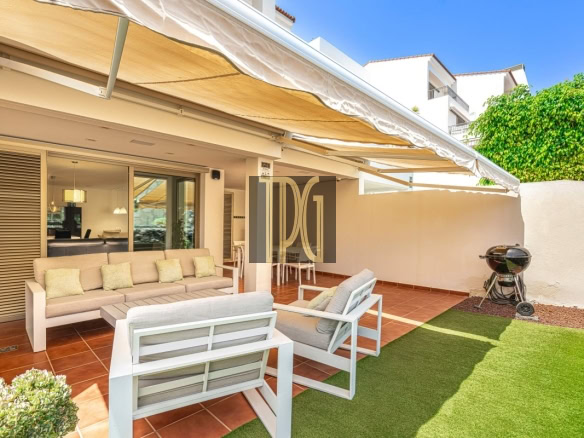 Patio lounge area of a modern home with beige outdoor sofa and white chairs under a retractable beige awning, adjacent to a grill and artificial turf.