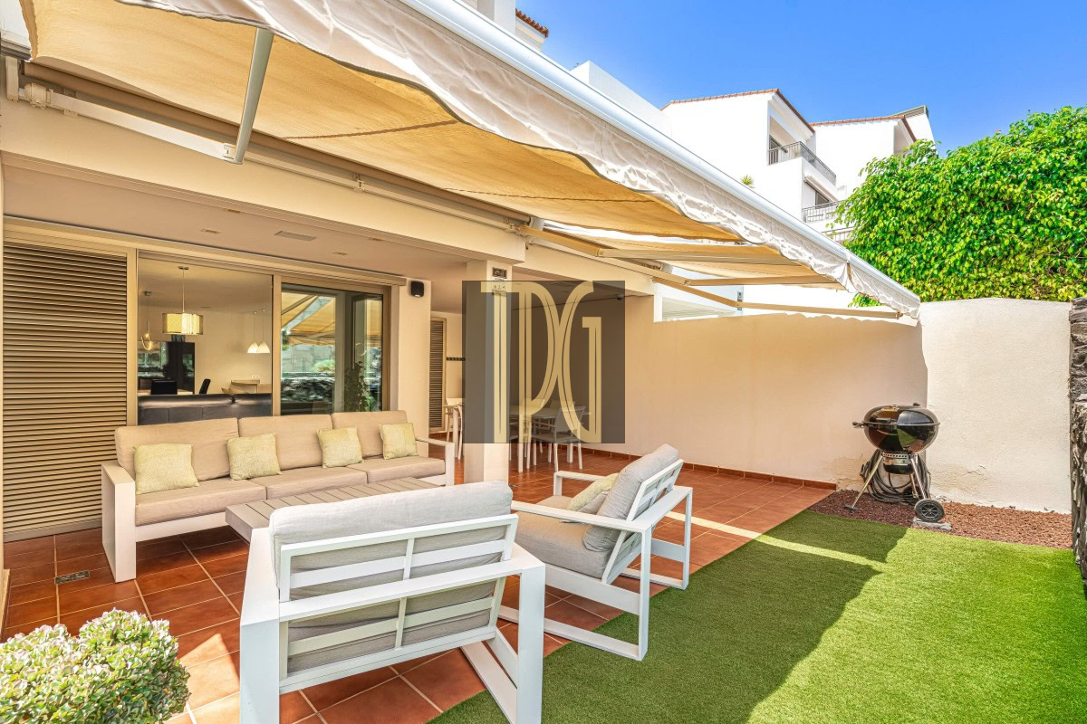 Patio lounge area of a modern home with beige outdoor sofa and white chairs under a retractable beige awning, adjacent to a grill and artificial turf.