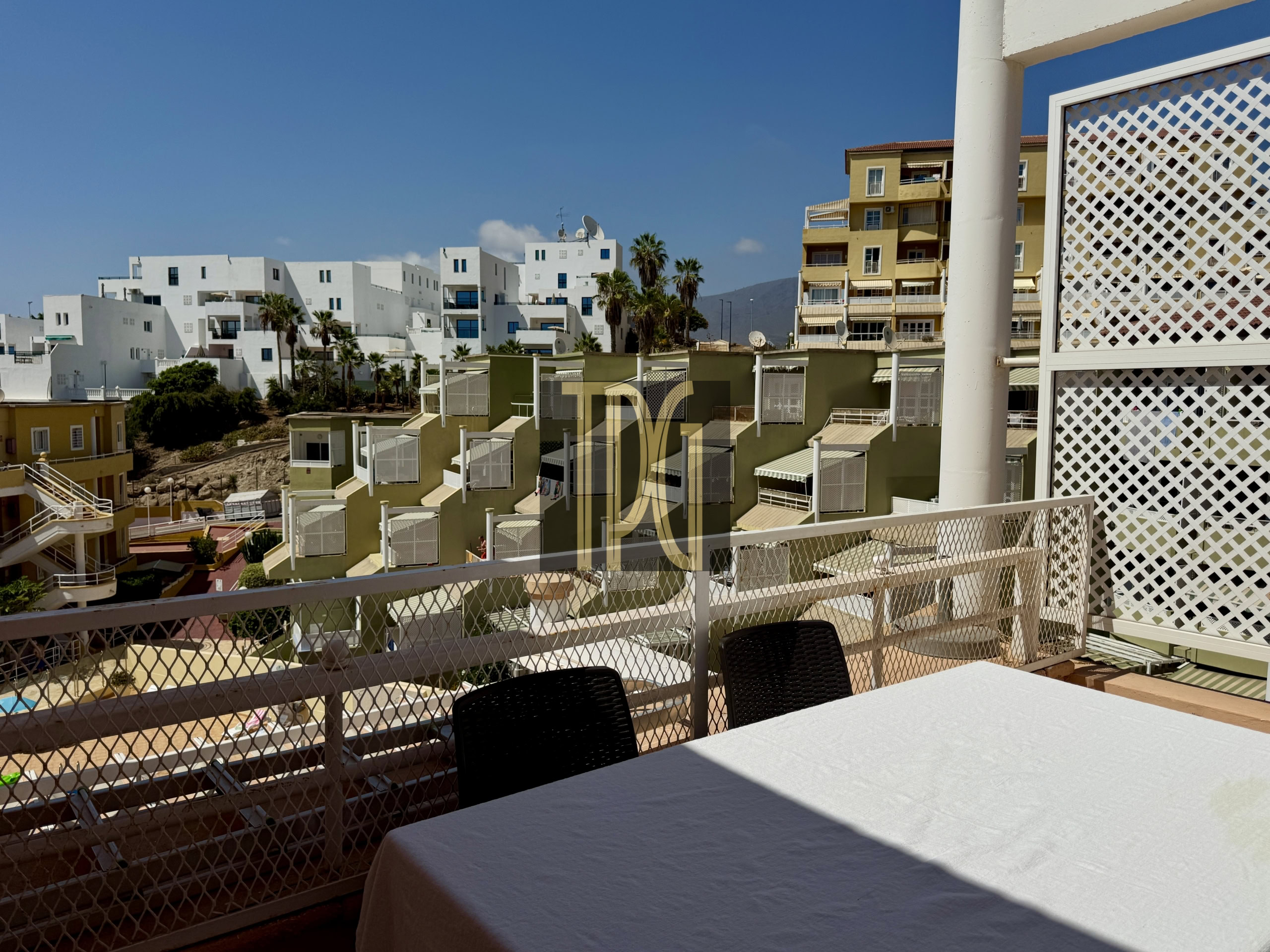 Outdoor terrace with a white-tablecloth table, black wicker chairs, and a lattice railing, overlooking white apartment buildings under a blue sky.