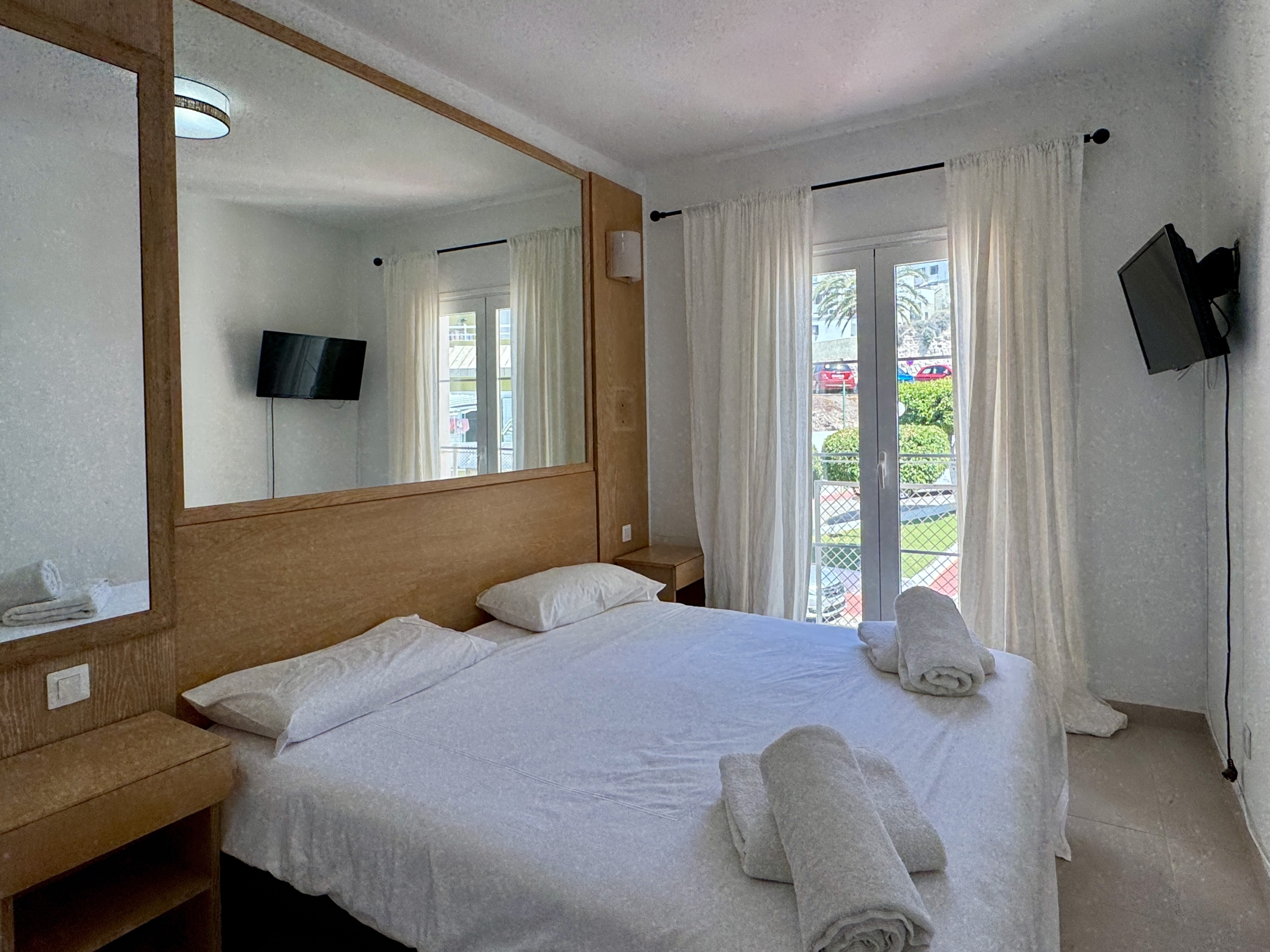 Hotel room with two double beds, white linens, folded towels, a large wooden headboard/mirror, and a wall-mounted TV on the side wall.