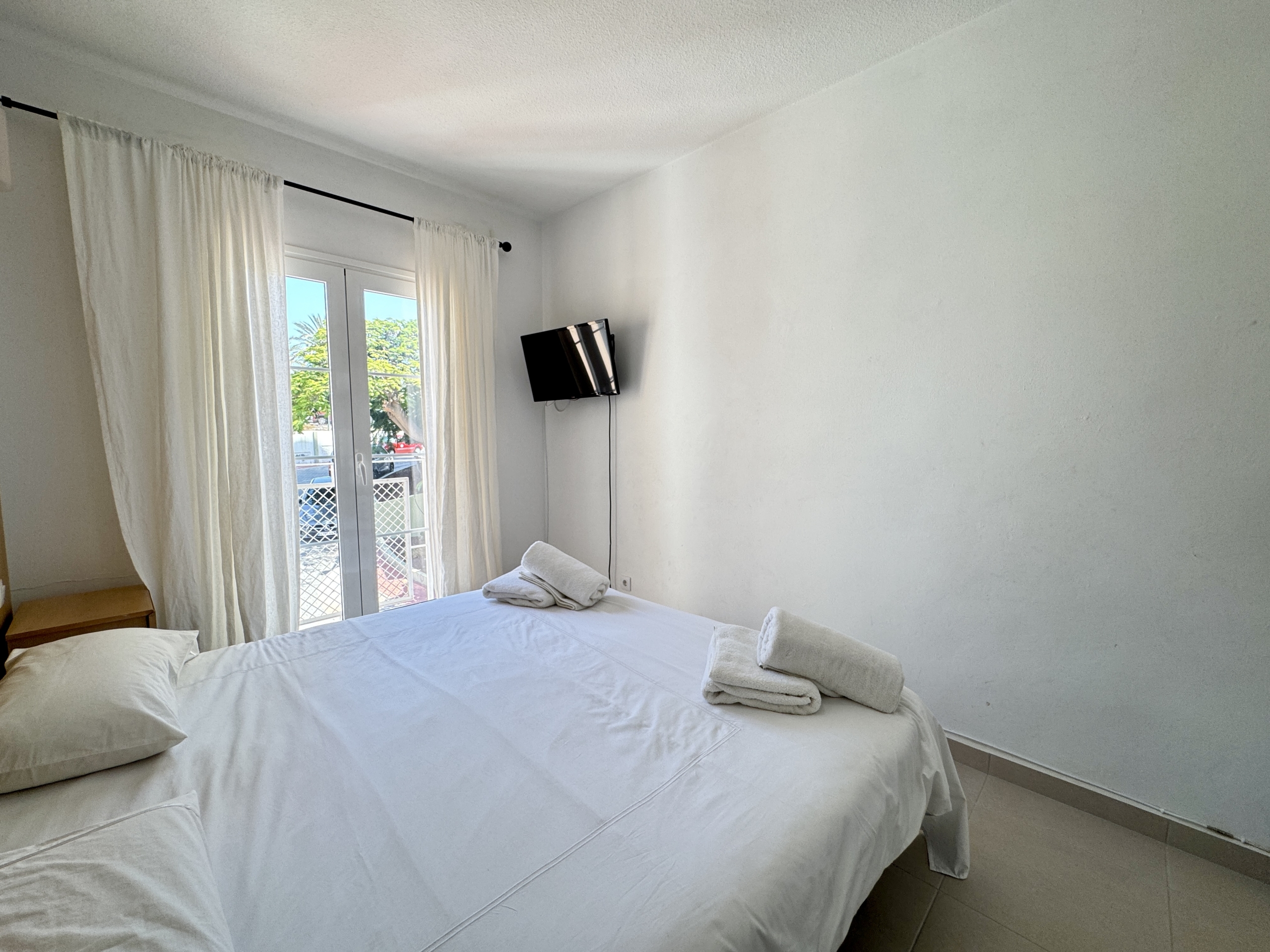 Twin beds with white linens and folded towels in a bright hotel room, French doors with white curtains, and a wall-mounted TV on the right wall.