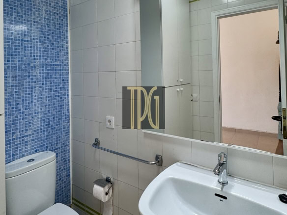 Bathroom with white sink and toilet, blue mosaic wall tile on left, mirror and cabinets above the sink.