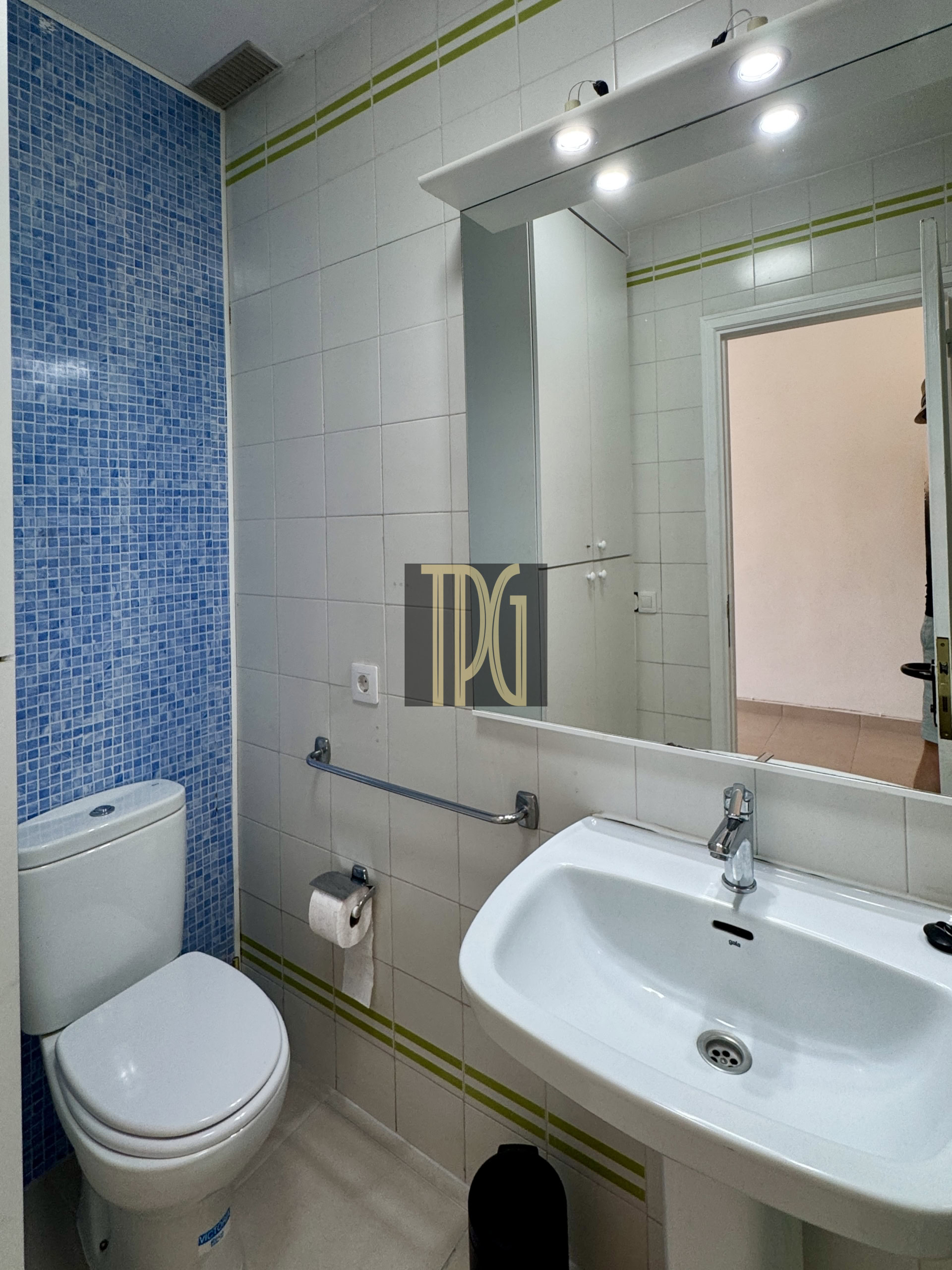 Bathroom with white sink and toilet, blue mosaic wall tile on left, mirror and cabinets above the sink.