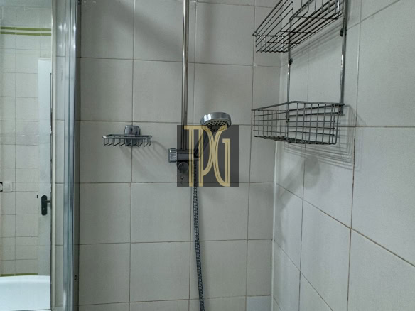 Shower area in a white-tiled bathroom with a vertical handheld shower, flexible hose, and two-tier chrome wire shelves on the wall, plus a glass door on the left side.