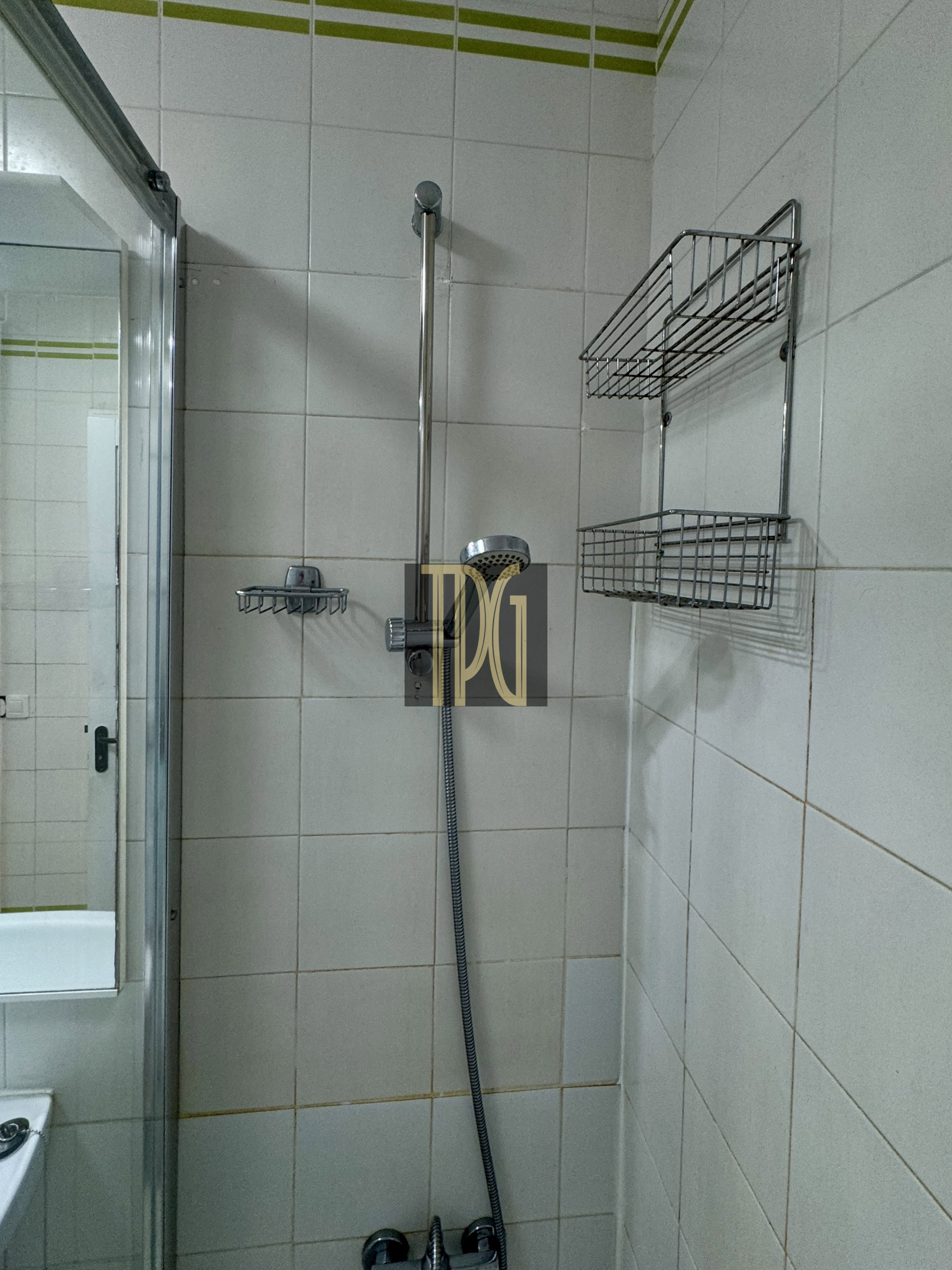 Shower area in a white-tiled bathroom with a vertical handheld shower, flexible hose, and two-tier chrome wire shelves on the wall, plus a glass door on the left side.