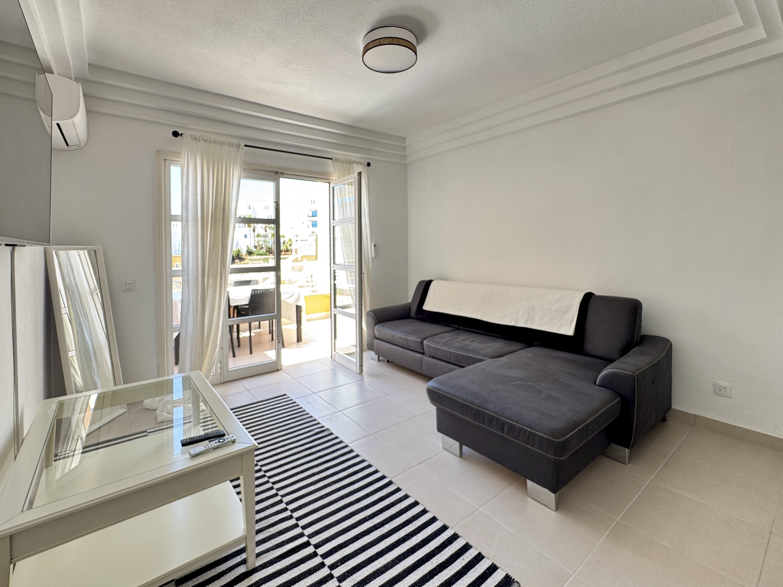 Bright living room with a dark L-shaped sofa, glass coffee table, and a black-and-white striped rug near open balcony doors.