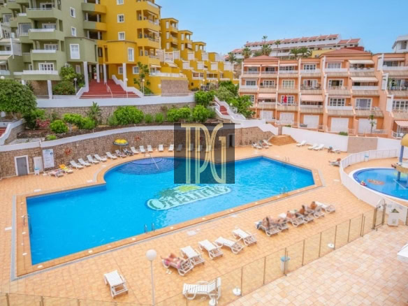 Resort-style swimming pool area with blue water, surrounded by orange-tiled deck and white sun loungers, and colorful apartment buildings in the background.