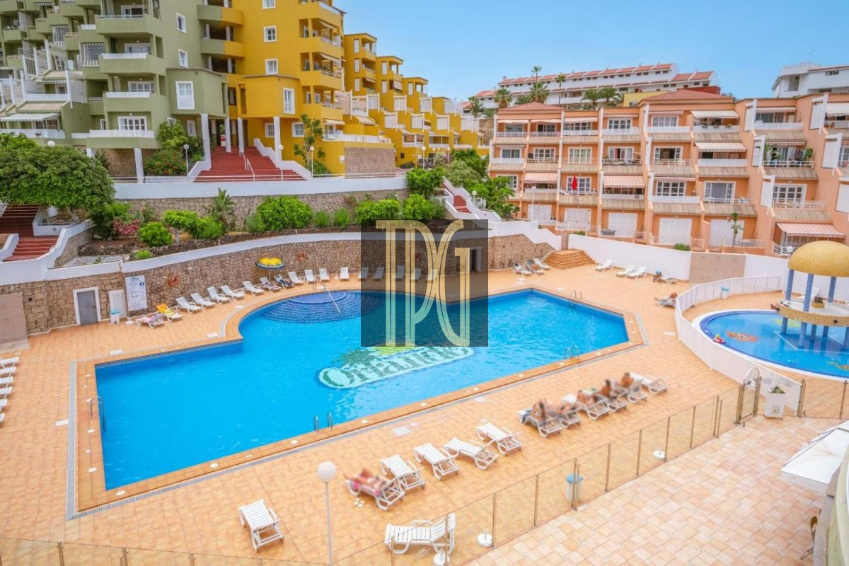 Resort-style swimming pool area with blue water, surrounded by orange-tiled deck and white sun loungers, and colorful apartment buildings in the background.