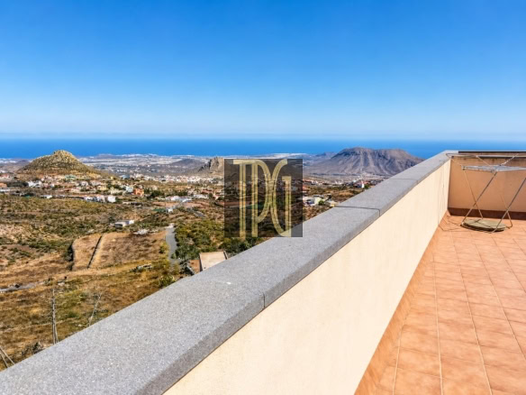 Terrace with tiled floor and low wall, overlooking arid hills, a small town, and the blue sea under a clear sky.