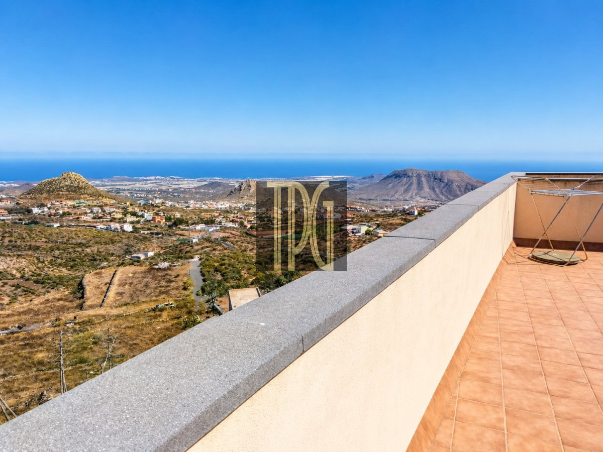 Terrace with tiled floor and low wall, overlooking arid hills, a small town, and the blue sea under a clear sky.