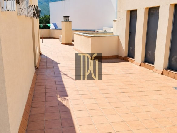 Sunlit rooftop terrace with terracotta tiles, beige walls, and a row of grey doors along the right; blue sky and distant mountains in the background.
