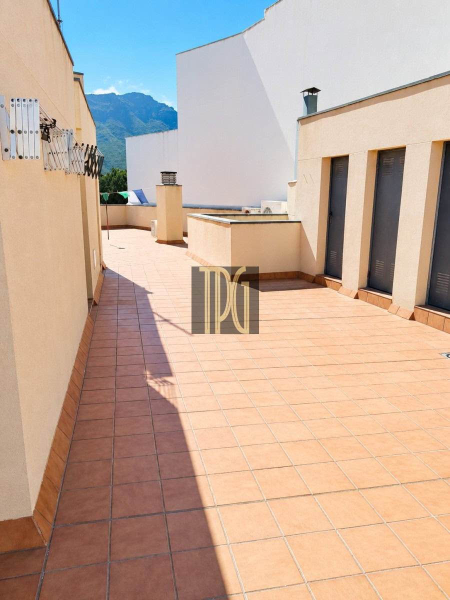 Sunlit rooftop terrace with terracotta tiles, beige walls, and a row of grey doors along the right; blue sky and distant mountains in the background.