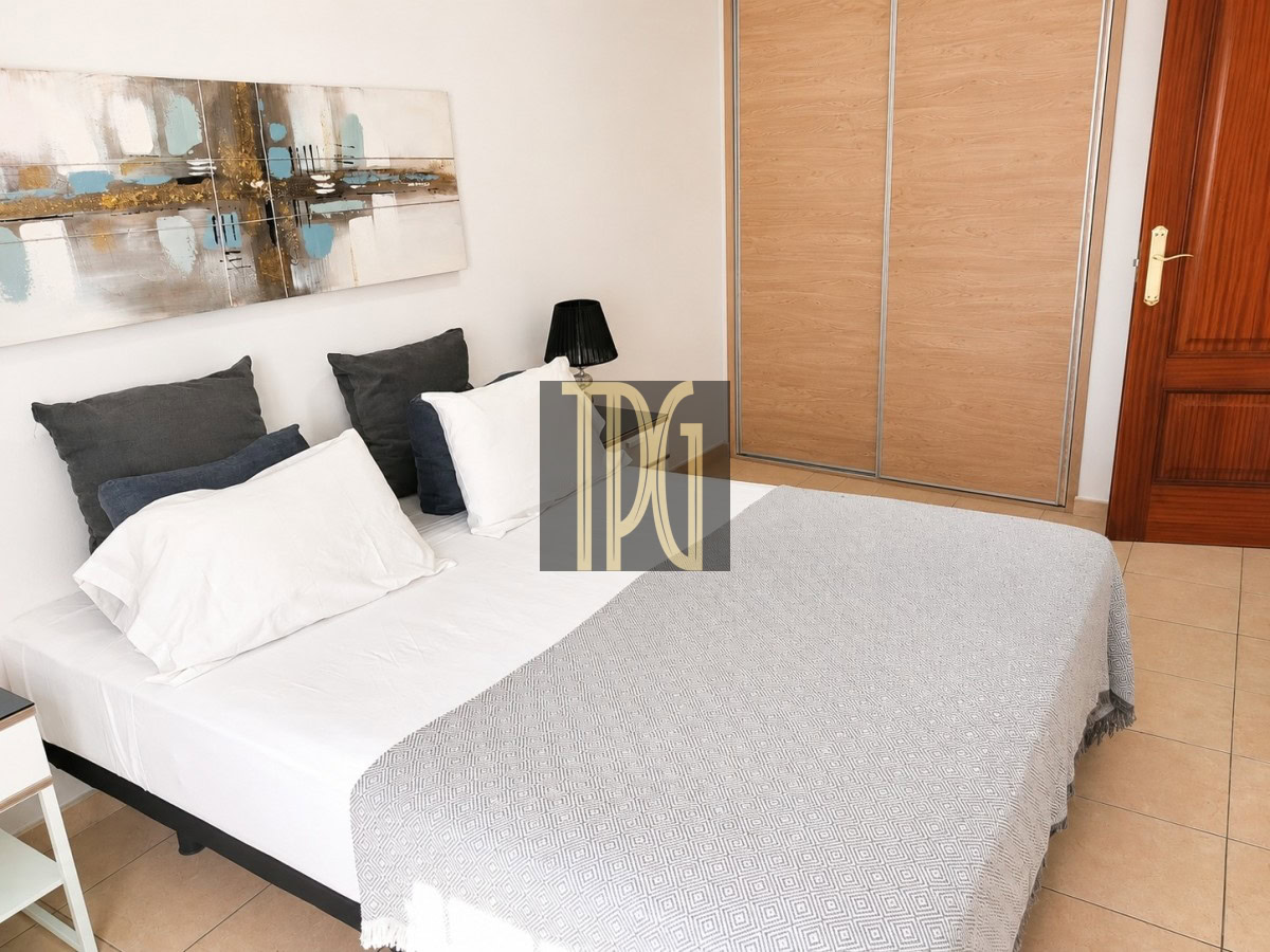 Modern bedroom with a made bed, white sheets, gray patterned throw, and multiple pillows, next to a black bedside lamp and abstract wall art.