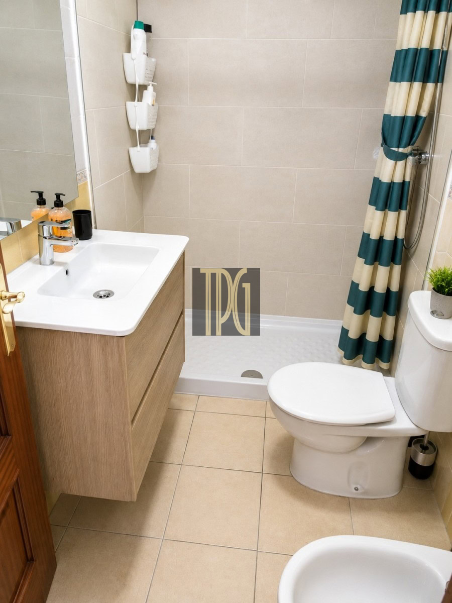 Small modern bathroom with a wooden vanity, white sink, and wall-mounted shower caddies beside a tiled shower area with a teal-striped curtain; toilet nearby.