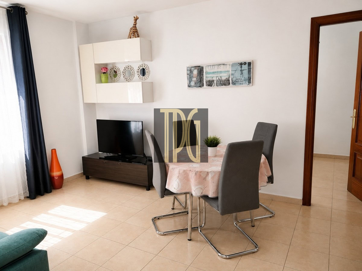 Living/dining area with round pink-dotted table and four gray upholstered chairs; TV on a dark wood stand against a white wall.