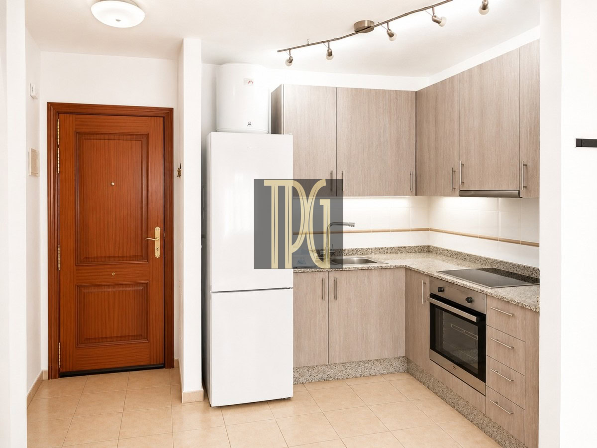 Compact L-shaped kitchen with light wood cabinets, white fridge, sink, and stainless steel oven under track lighting in a small galley space.