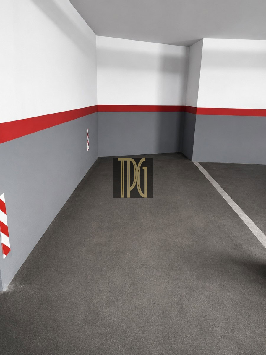 Empty parking space in a concrete garage with white walls above, a red stripe across the middle, and gray lower walls; a white line marks the next spot edges and a black logo sits on the floor.