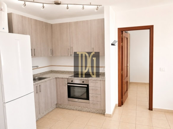 Light-filled L-shaped kitchen with light wood cabinets, granite countertops, a built-in stainless steel oven, and a sink; white fridge on the left and tiled floor; doorway with a wooden frame opens to a hallway on the right.