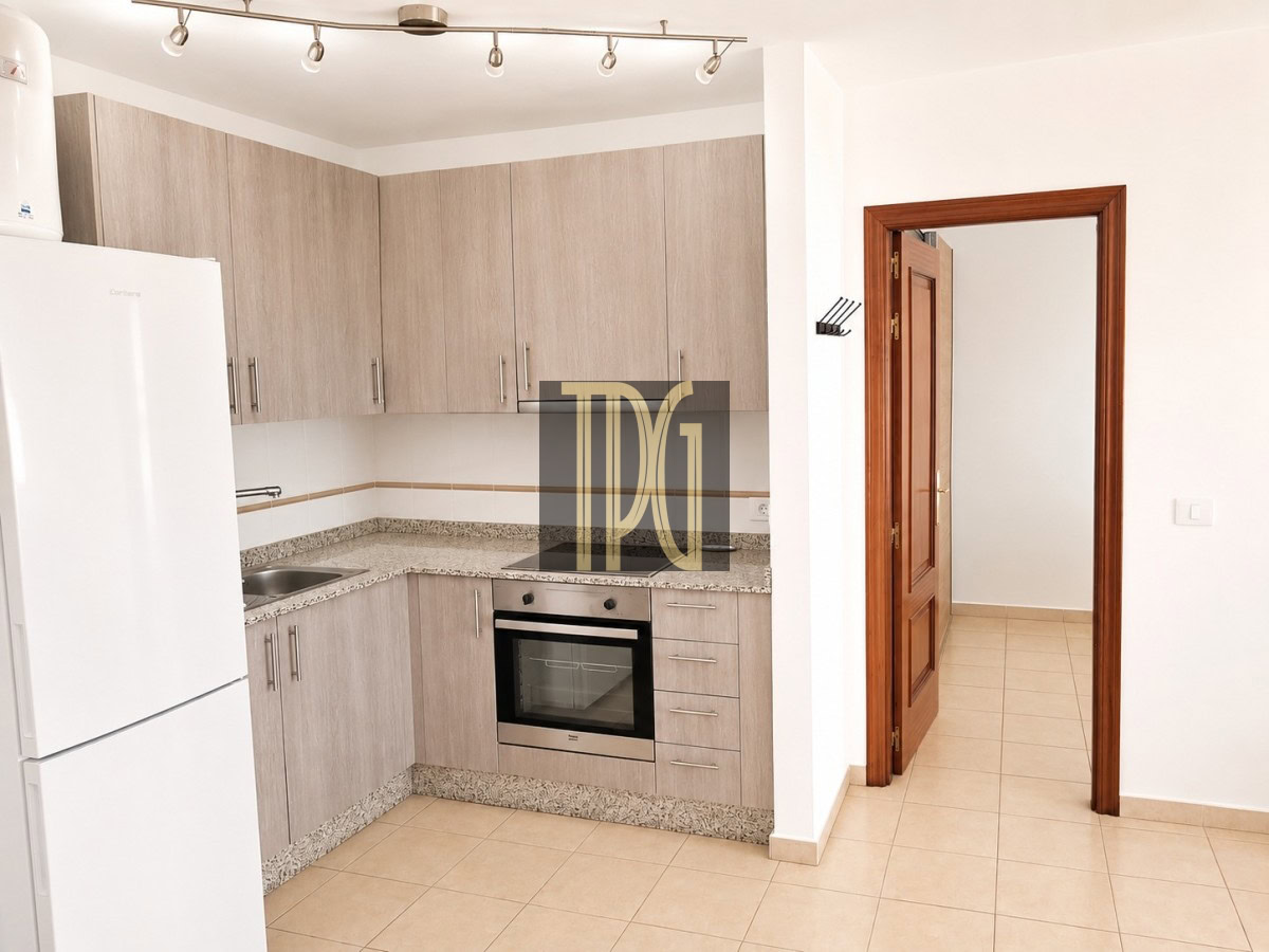 Light-filled L-shaped kitchen with light wood cabinets, granite countertops, a built-in stainless steel oven, and a sink; white fridge on the left and tiled floor; doorway with a wooden frame opens to a hallway on the right.