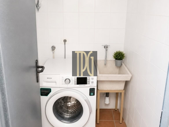 Small laundry nook with a front-loading washing machine next to a beige utility sink and a potted plant on a wooden stand, in a white-tiled room with terracotta floor tiles.