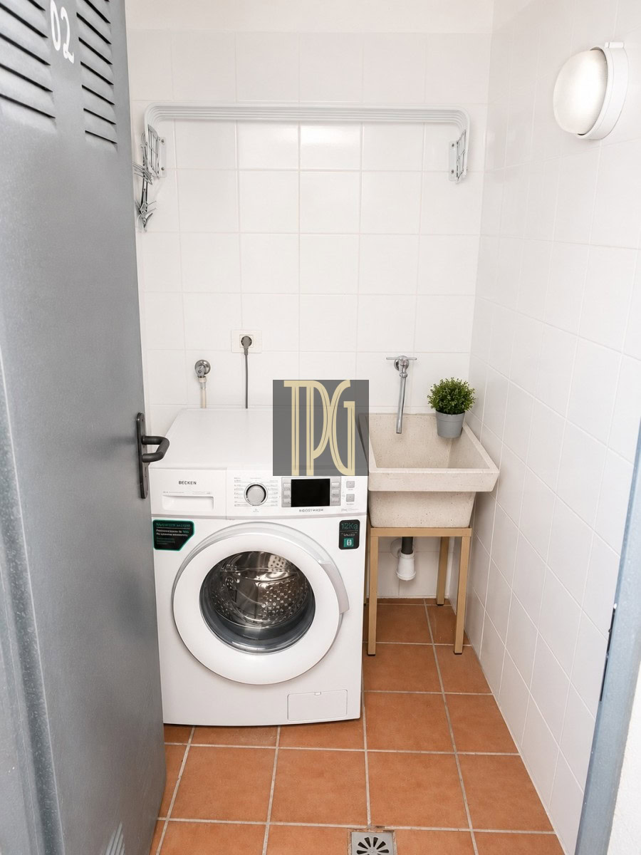 Small laundry nook with a front-loading washing machine next to a beige utility sink and a potted plant on a wooden stand, in a white-tiled room with terracotta floor tiles.