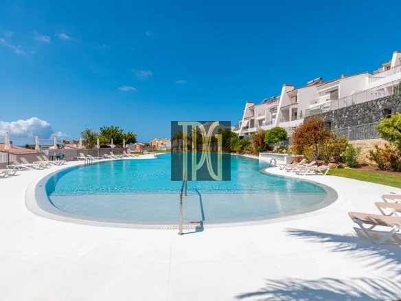 Resort swimming pool surrounded by white lounge chairs under a blue sky with modern white buildings and greenery nearby