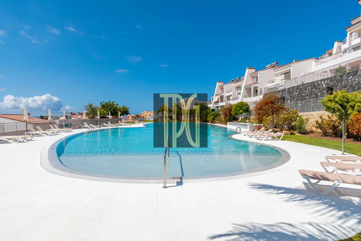 Resort swimming pool surrounded by white lounge chairs under a blue sky with modern white buildings and greenery nearby
