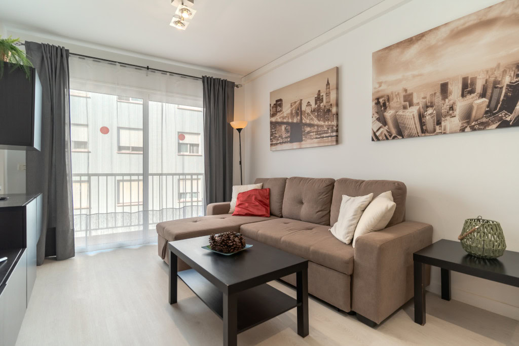 Cozy living room with a tan sofa, white and red cushions, black coffee table, and city skyline art on the wall.