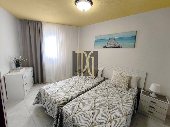Twin bedroom with two single beds, white headboard and matching nightstands, gray-patterned quilts, and a coastal artwork above the bed window.