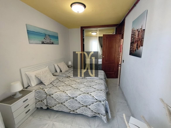 Bright hotel room with a large bed, two white nightstands with lamps, beach-themed art on the left, and an open wooden door leading to another room.