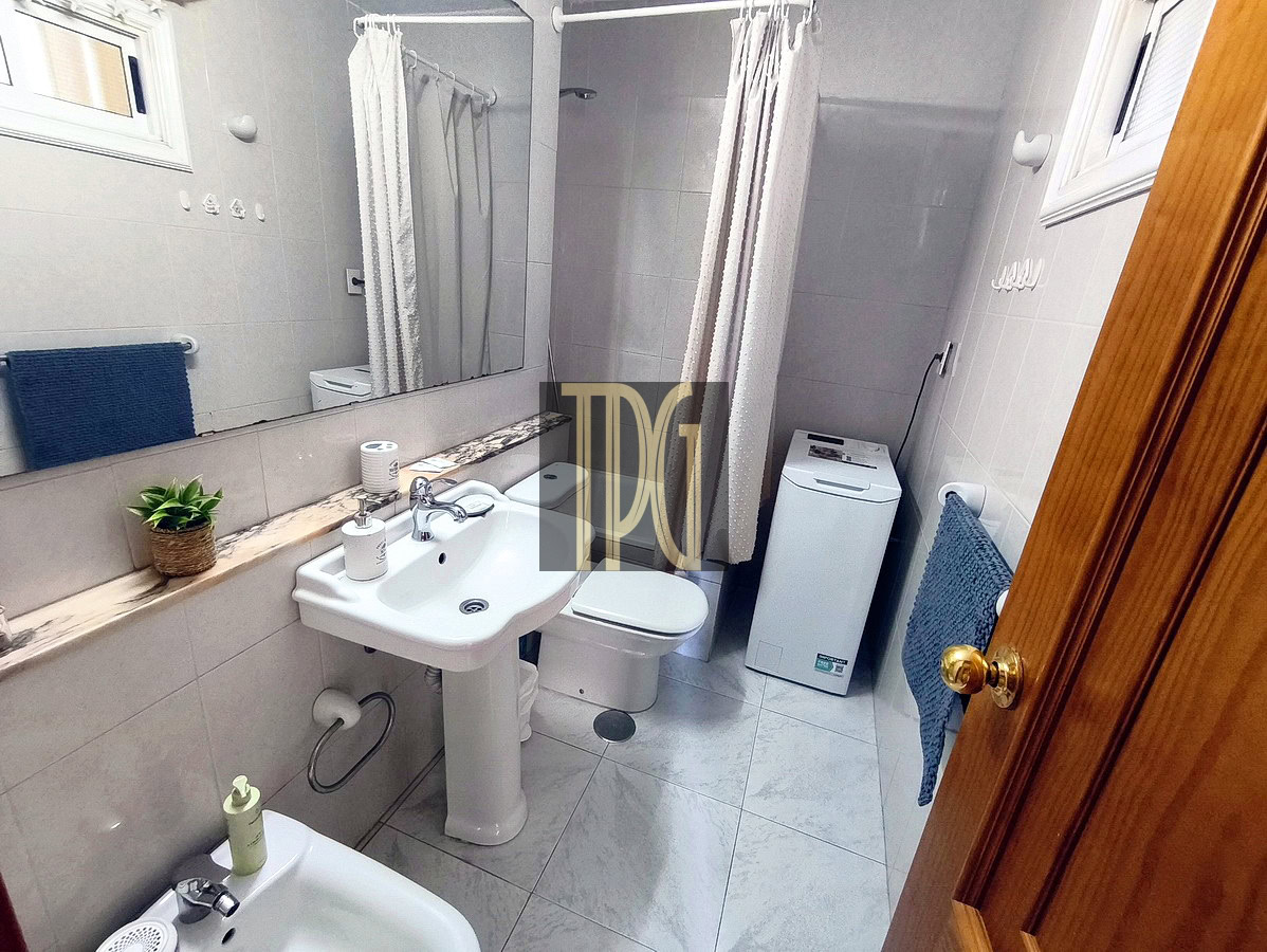 Small tiled bathroom with pedestal sink, toilet, washing machine, and a shower behind a white curtain; blue towels and a potted plant on a marble ledge.