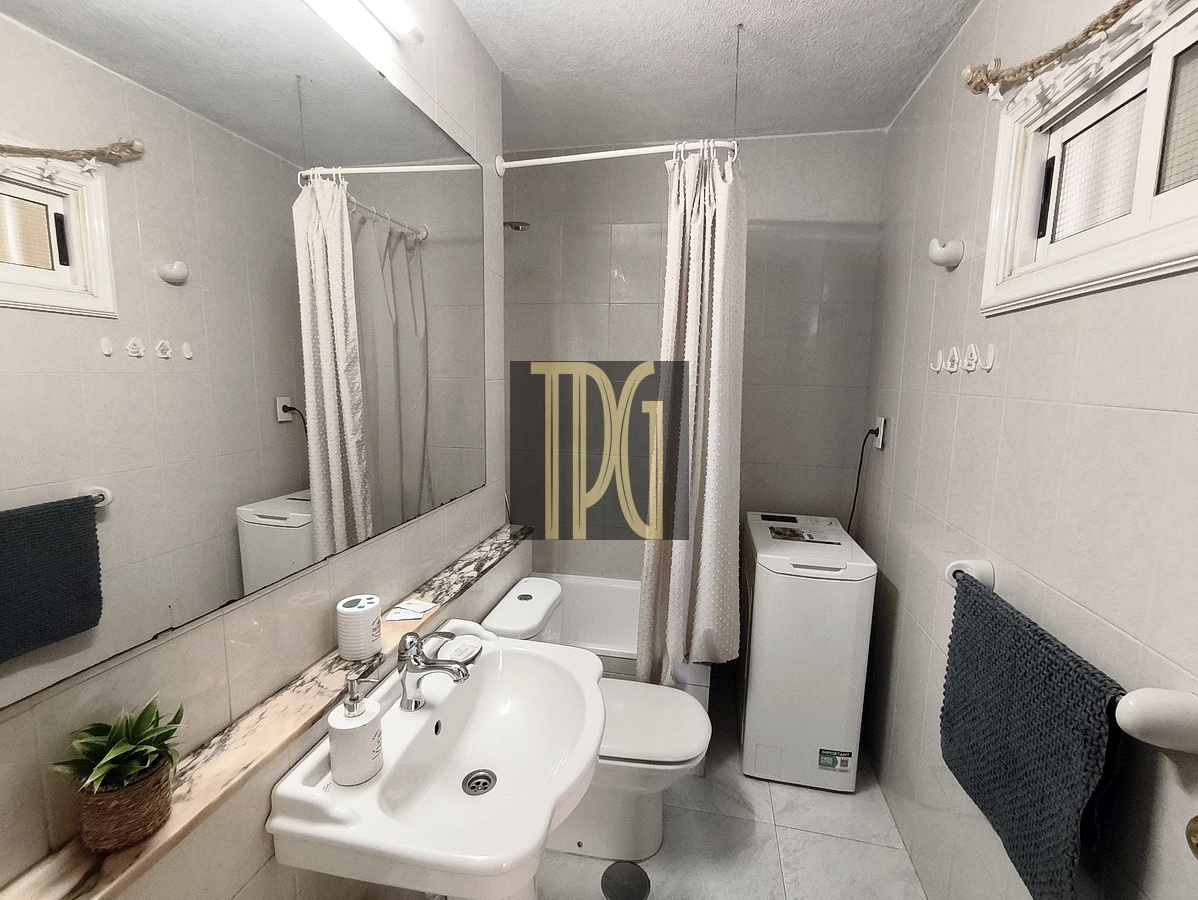 Small white bathroom with sink, toilet, washing machine, and a shower with a curtain along a marble counter and mirrored wall