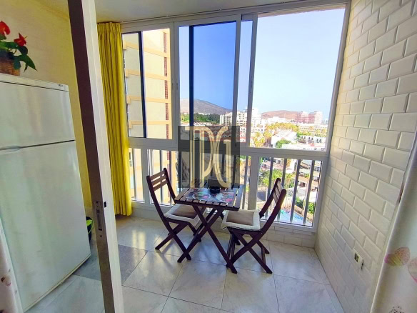 Enclosed balcony with a small folding table and two chairs, overlooking a city skyline and hills; refrigerator on the left and yellow curtains nearby.